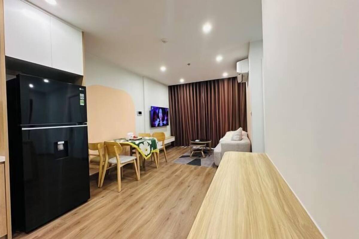Una habitación de hotel moderna con piso de madera, un refrigerador negro, una mesa de comedor con sillas, un sofá y un televisor en la pared.
