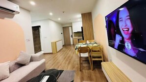 A modern living room with a large flat-screen TV, a sofa, and a dining area with wooden chairs and a table. - 2BRS@Vinhomes Smart City (Hanoi)