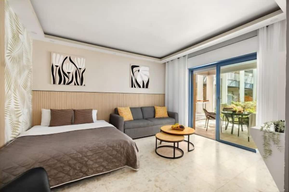 A modern hotel room with a bed, sofa, and a small table. There are abstract wall art pieces and a view of the outdoors through a glass door.