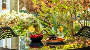 A table with a glass top, surrounded by chairs, in a garden setting with various plants and flowers.