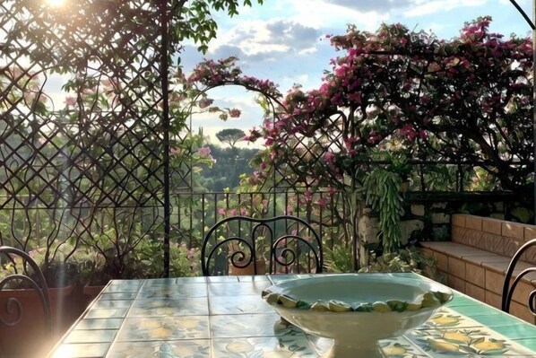 Une table avec un bol, une chaise et un banc, le tout installé dans un jardin, avec une clôture en métal et des plantes en fleurs.