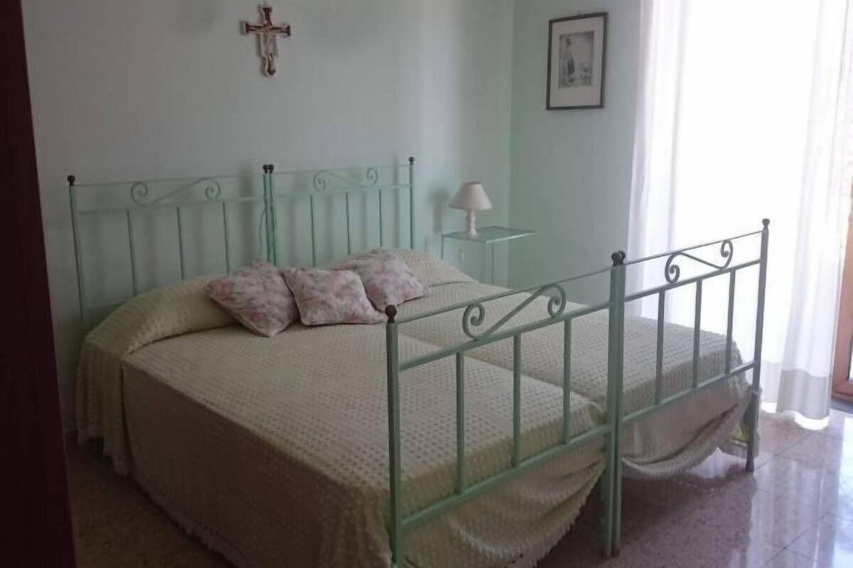 A single bed with a metal frame, a bedside table with a lamp, and a cross on the wall.