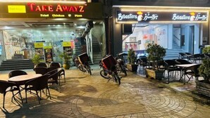 An outdoor seating area with tables and chairs, a bicycle, and a storefront with signs for 'TAKE AWAYZ' and 'Broasters'. - Premium Apartments (Gujranwala)