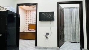 A compact room with a bed, a TV mounted on the wall, and a refrigerator. - Premium Apartments (Gujranwala)