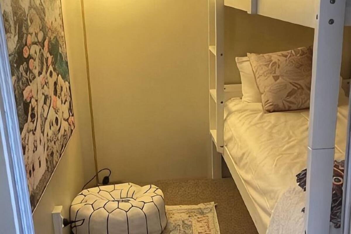 Una cama individual doble con un cojín estampado, un otomanillo redondo y una pared con un cuadro floral.