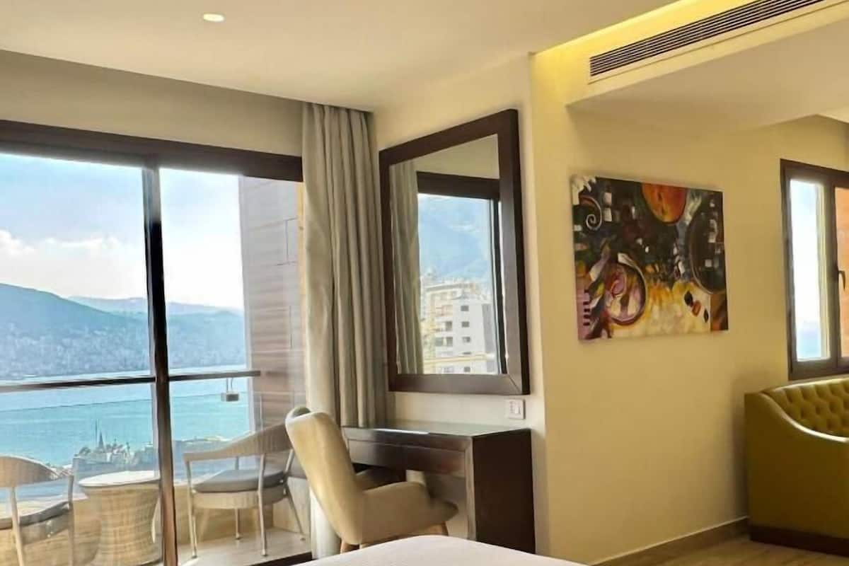 Habitació d'hotel amb un llit gran, una taula de treball, una cadira, un quadre i vista al mar.