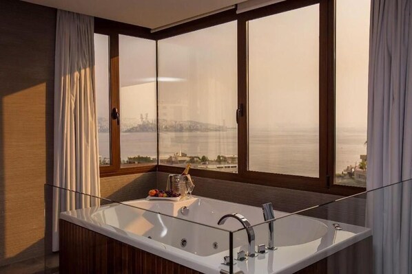 A modern bathroom with a large bathtub, a glass-enclosed shower, and a view of a cityscape through large windows. - OPERA HOTEL (Kfar Yassine)