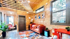 Ein gemütliches Blockhaus-Interieur mit Holzbalkendecke, einer Couch, einem Esstisch und einem großen Fenster mit Blick auf einen verschneiten Wald.