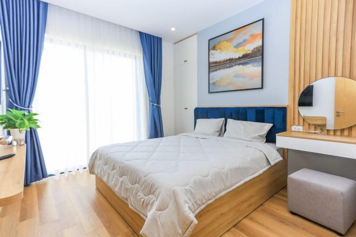 A modern bedroom with a large bed, a bedside table, a mirror, and a view of the outdoors.