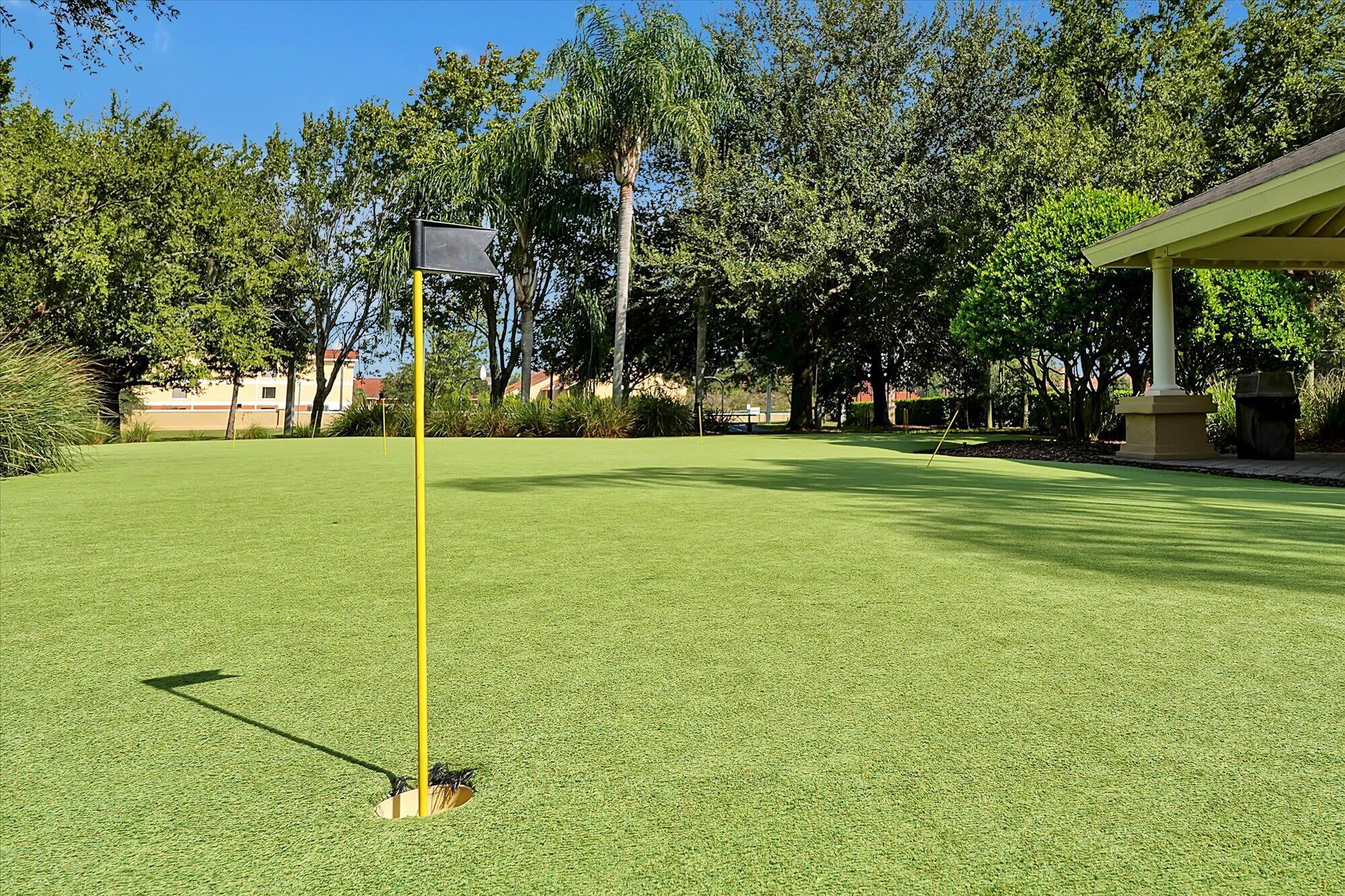 A golf course with a well-maintained green, a yellow flagstick, and a building in the background.