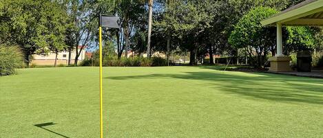 A golf course with a well-maintained green, a yellow flagstick, and a building in the background.