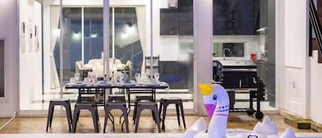 An indoor swimming pool with inflatable toys, a dining area with a table and chairs, and a view through glass doors.