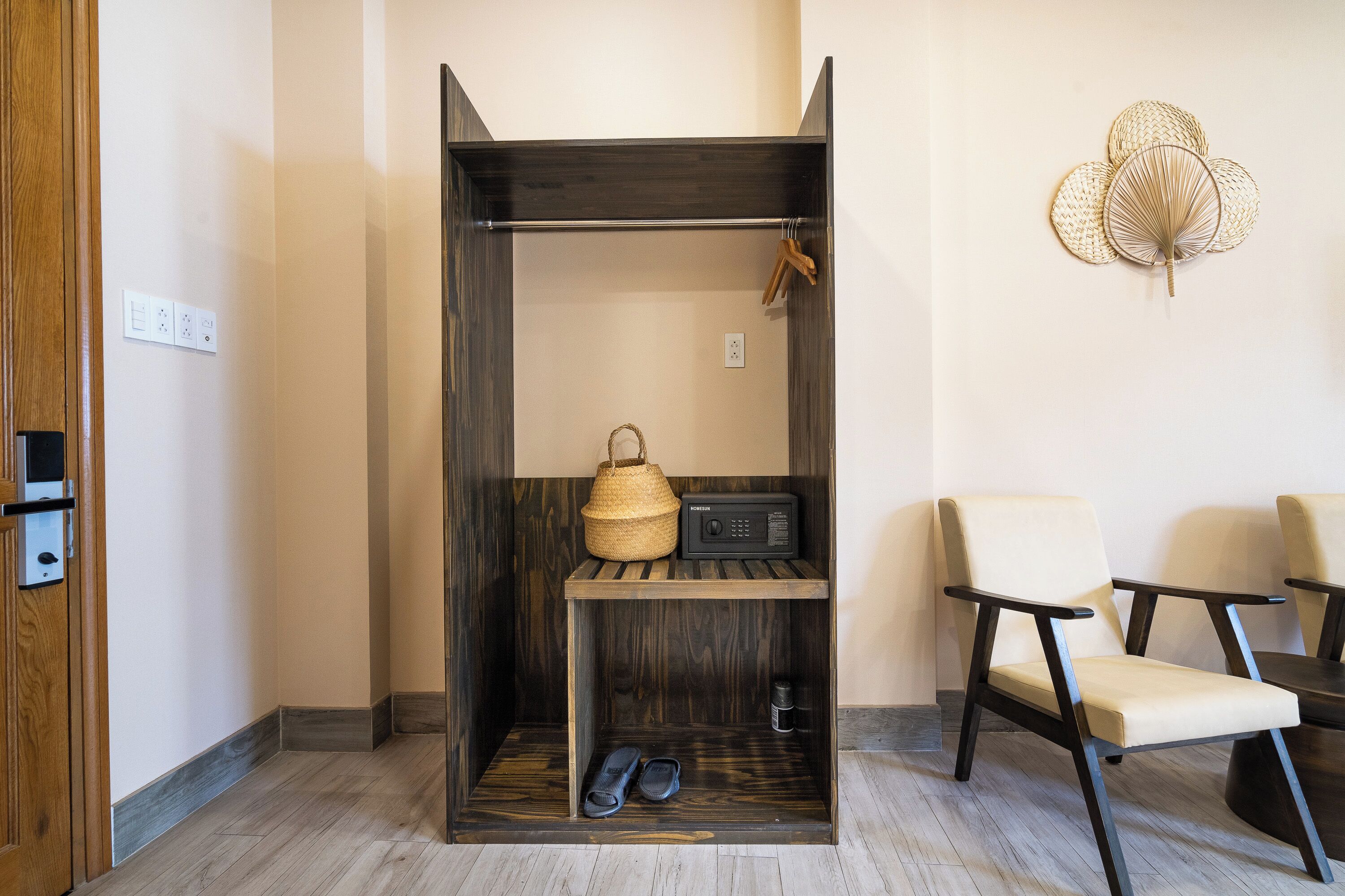 A compact room with a wooden wardrobe, a shelf with a woven basket and a radio, a pair of slippers, and two chairs.