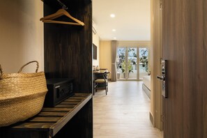 Habitación de hotel con piso de madera, una cesta grande de fibra natural, un dispositivo electrónico negro, un banco de madera, una mesa de comedor con sillas y un ventanal corredero que da a un balcón con plantas.