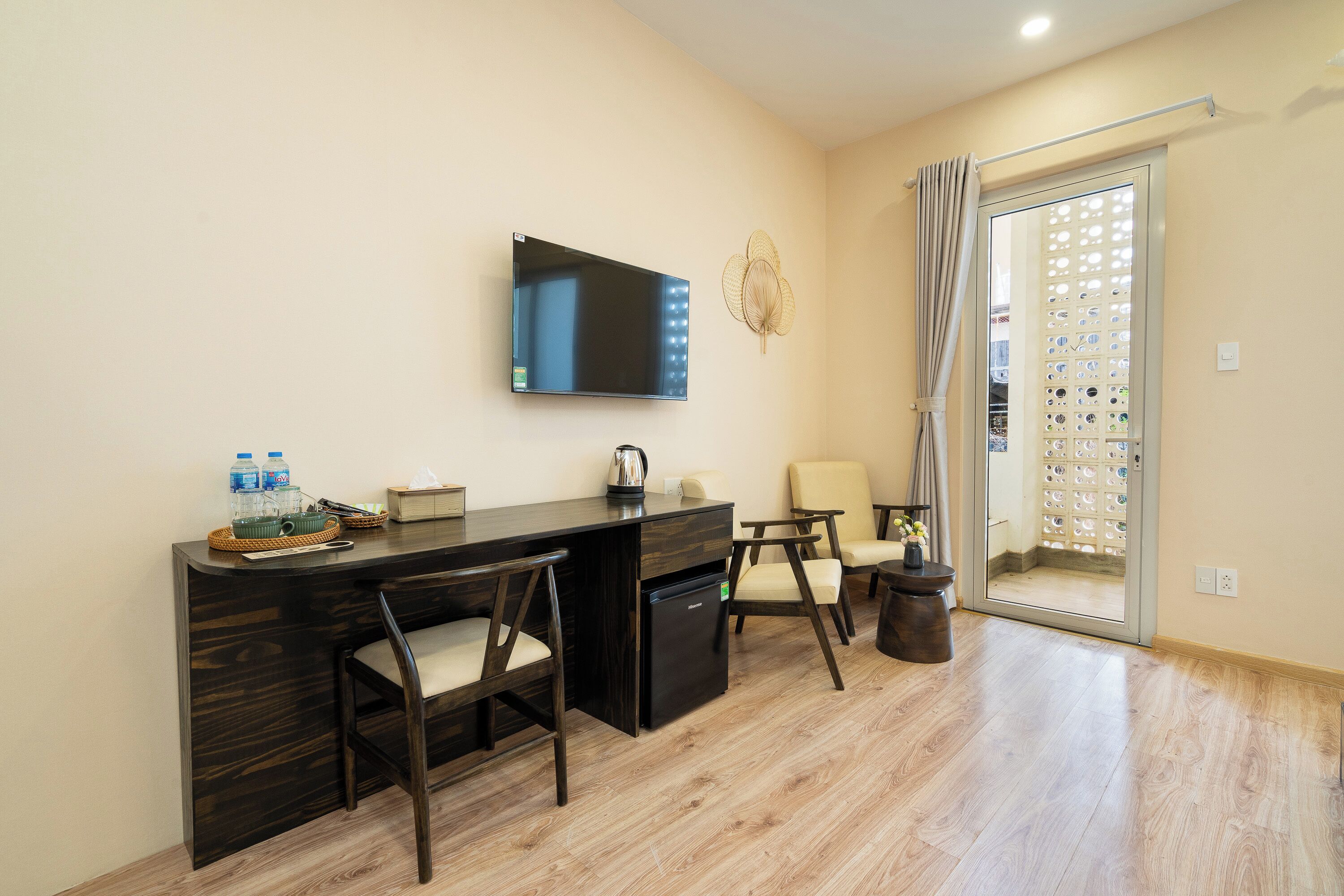 A modern hotel room with a dark wood desk, two chairs, a flat-screen TV, and a sliding glass door leading to a balcony.