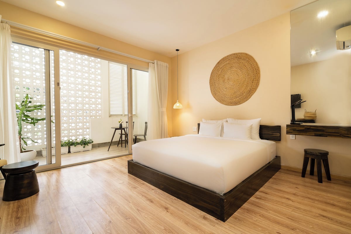 Un dormitorio con una cama grande, un taburete pequeño, un adorno circular tejido en la pared y una puerta corrediza de vidrio que da a un balcón con plantas.