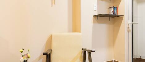 A room with a beige chair, a small round table with a vase of flowers, a wall-mounted decorative fan, and a shelf with books and a bottle.