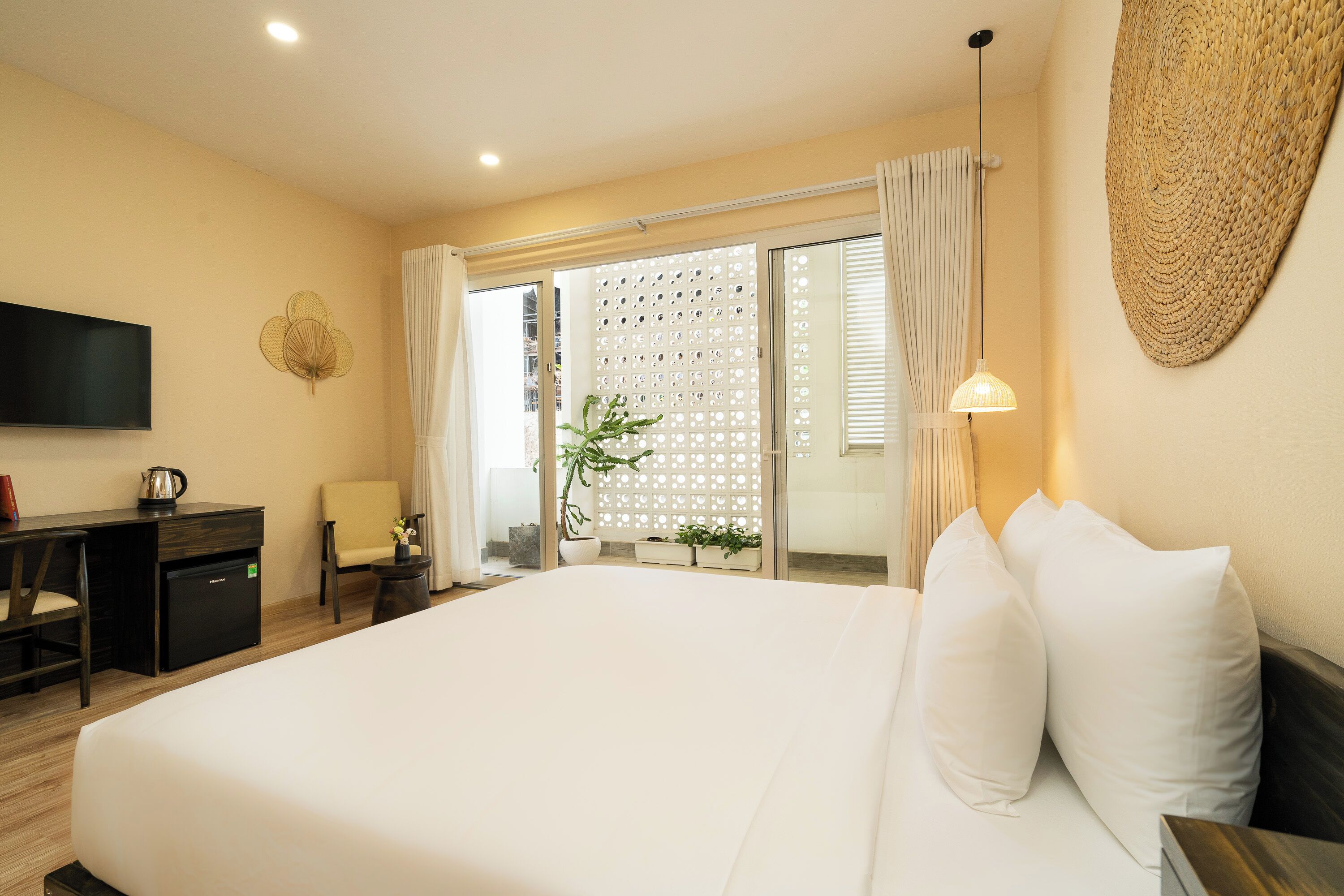 A hotel room with a large bed, a desk with a TV, a small table, and a potted plant.