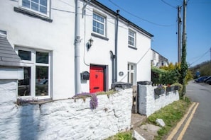 A white building with a red door, stone wall, and a small garden area with flowers. - Rose Cottage - 3 Bedroom Cottage - Parkmill (Ilston)