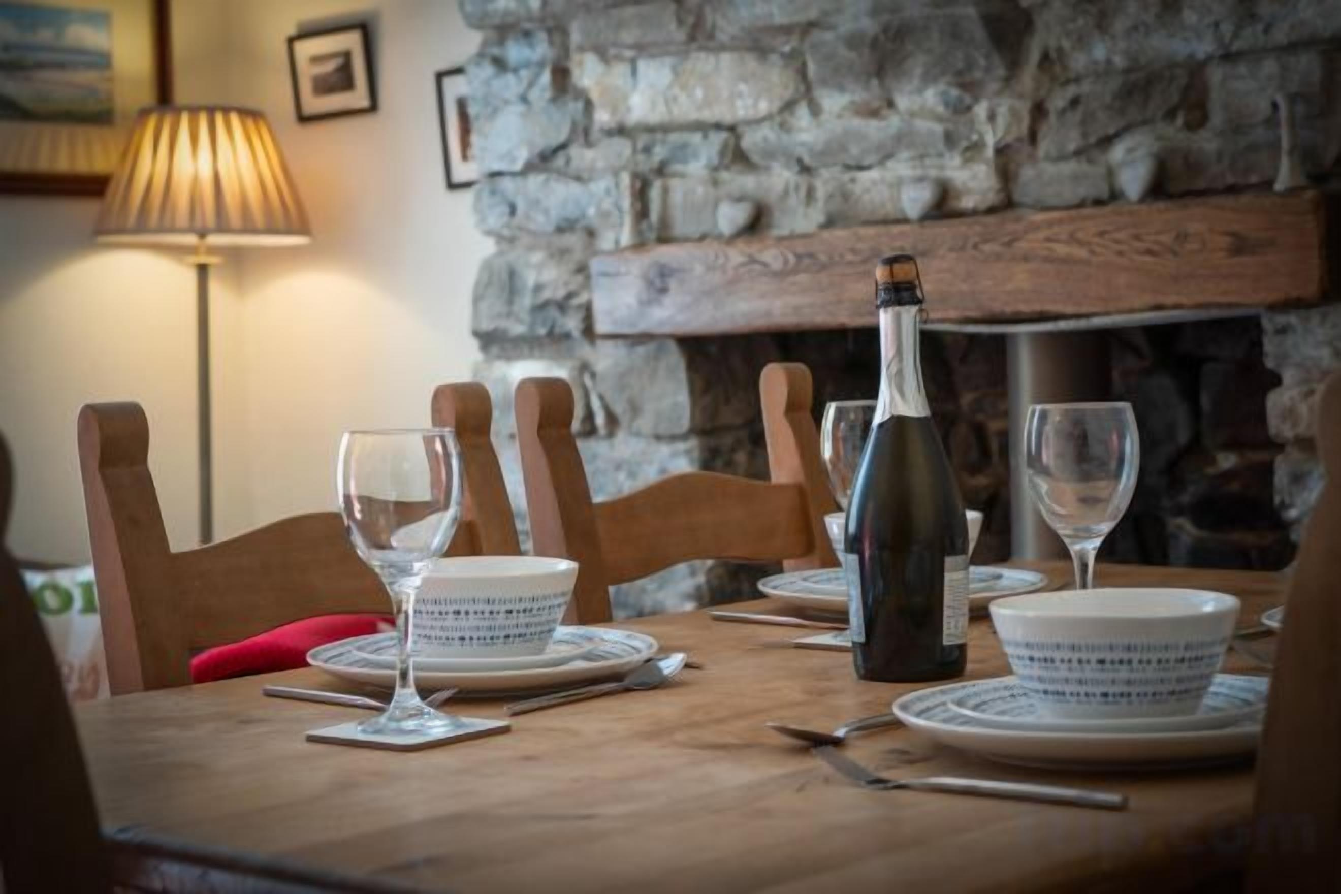 A dining table set with plates, glasses, and a bottle, with a stone wall and fireplace in the background.