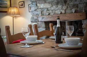 Mesa lista con platos, vasos y una botella, con una pared de piedra y chimenea al fondo.