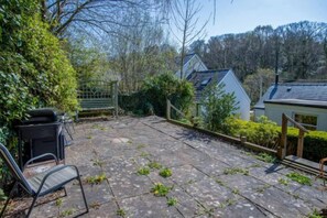 A paved patio area with a metal chair and a table, surrounded by greenery and a wooden fence. - Rose Cottage - 3 Bedroom Cottage - Parkmill (Ilston)