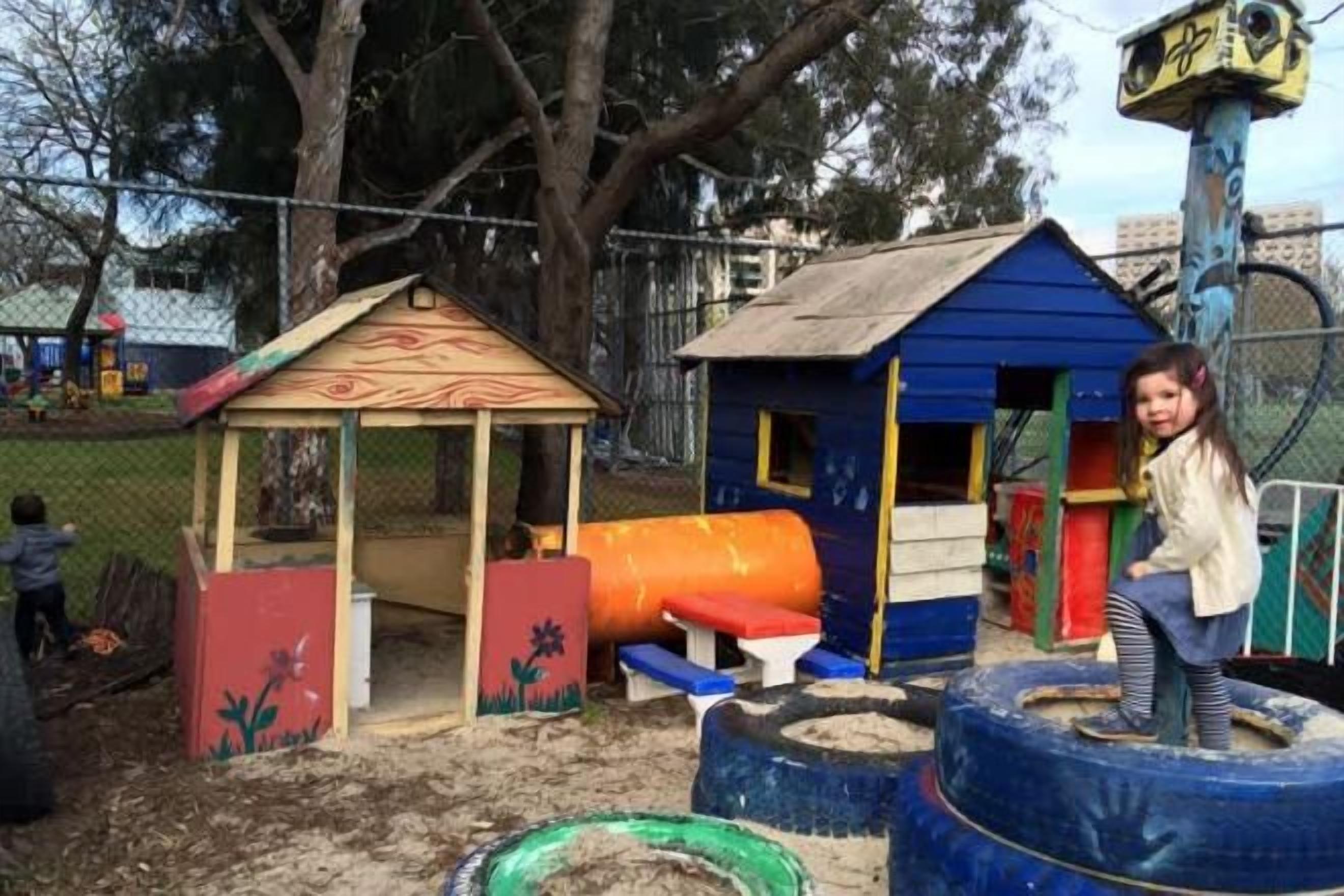 Un parque infantil con una casita azul, un tobogán y una zona de juegos con neumáticos.