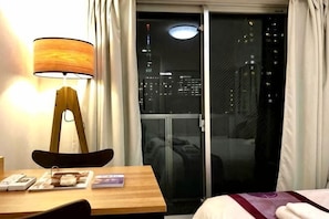 A hotel room with a wooden table, a lamp, a chair, a bed, a window with curtains, and a view of the city at night. - Kinshicho 1001 (Tokyo)