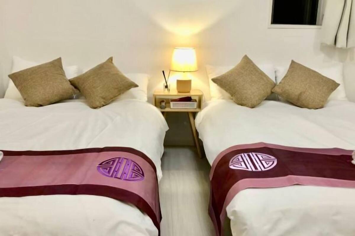 A hotel room with two beds, each with a pillow and a blanket. There is a nightstand with a lamp between the beds.