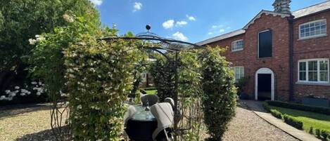 Een tuin met een prieel, een gebouw met een bakstenen gevel en een grindpad.