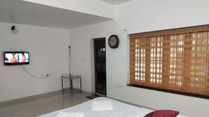 A hotel room with a bed, a television, a clock, and a window with blinds. - Azza Heritage Home (Kochi)