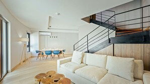 Un salon moderne avec un canapé blanc, un coin repas avec des chaises bleues et un escalier doté d’une rampe noire.