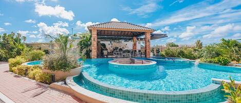 A swimming pool with a sunken hot tub, surrounded by a covered seating area and lush landscaping.
