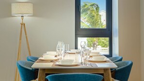 Une table à manger est dressée avec des assiettes, des verres et des couverts, placée devant une fenêtre qui offre une vue sur des palmiers et des maisons.