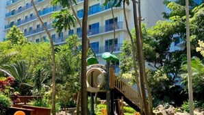 Un hotel de varios pisos con balcones y un exuberante jardín con un parque infantil.