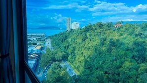A view from a window overlooking a cityscape with a road, buildings, and a hill with dense greenery. - JQ Central 4 person unit facing mountain view and partially seaview AT kk CITY CENTER (Kota Kinabalu)