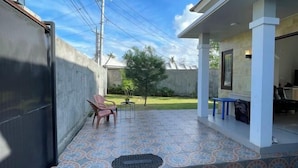 A covered patio area with a patterned floor, a small seating arrangement, and a view of a residential area in the background. - Breeze House (West Lombok Regency)