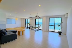 A spacious living area with a sofa, a dining table, a hammock, and large windows with a view of the sea.