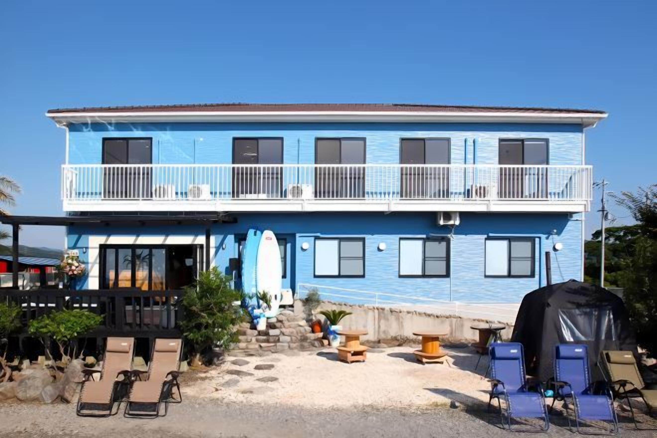 Un edificio de dos plantas con un balcón, mobiliario exterior y una decoración con una tabla de surf.