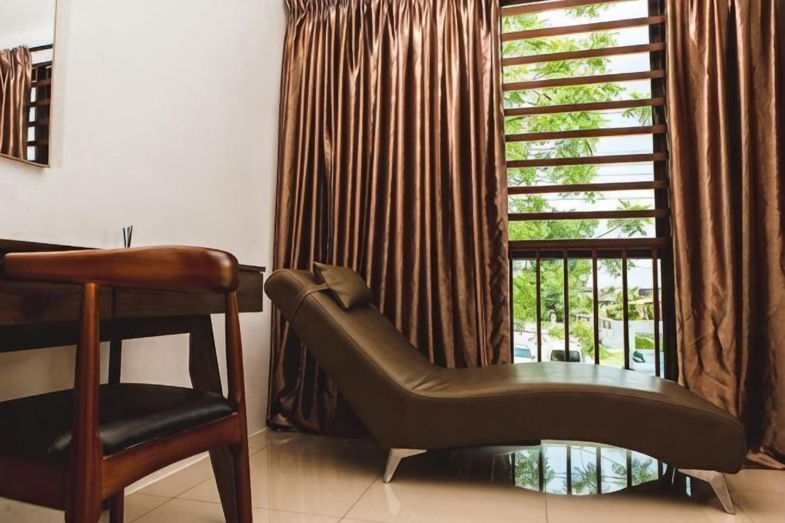 A room with a curved lounge chair, a wooden chair, and a window with vertical blinds.