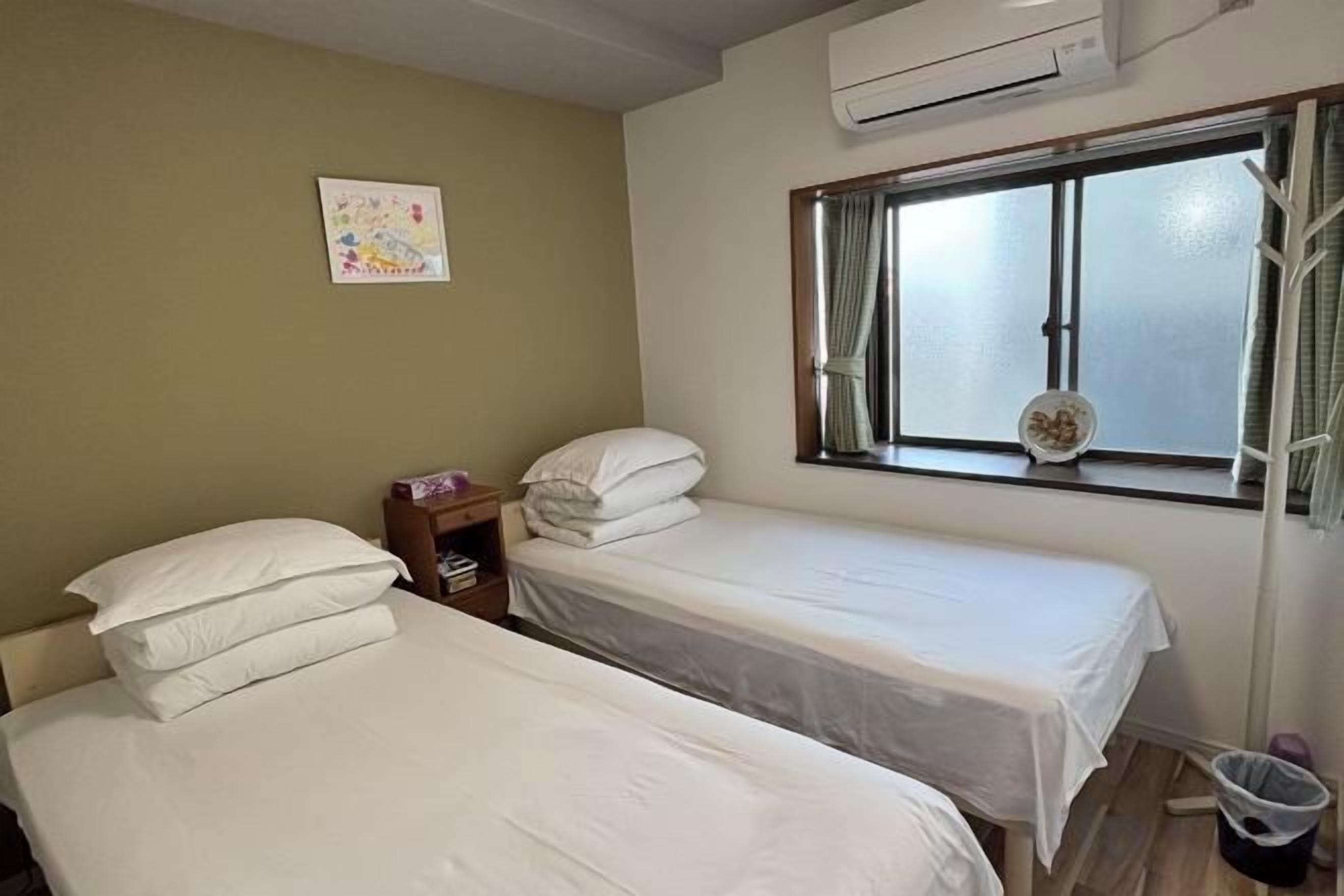 A room with two single beds, a small bedside table, a window with curtains, and a wall-mounted air conditioner.
