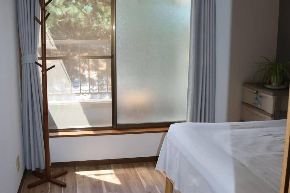 A bedroom with a bed, a window with curtains, a wooden floor, and a plant on a side table.