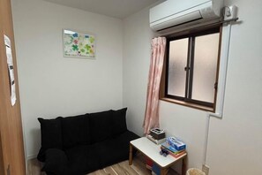 A small, sparsely furnished room with a black sofa, a small table with books and a trash bin, a window with a pink curtain, and an air conditioning unit mounted on the wall. - XUYUAN (Osaka)