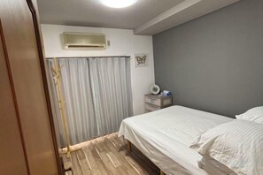 A bedroom with a bed, a bedside table, an air conditioning unit, a curtain, and a wall-mounted light fixture. - XUYUAN (Osaka)