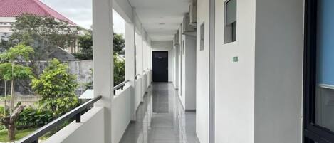 Un couloir avec une rambarde, une porte au fond, et une vue sur un bâtiment avec un toit rouge.