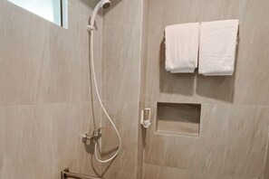 A shower area with a handheld showerhead, a towel rack with two towels, and a small shelf. - Puri Residence Hotel (Sananwetan)