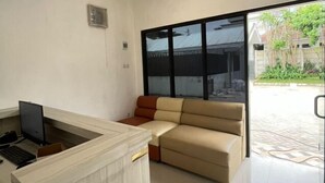 A modern living room with a sofa, a desk with a computer, and a large sliding glass door leading to an outdoor area. - Puri Residence Hotel (Sananwetan)