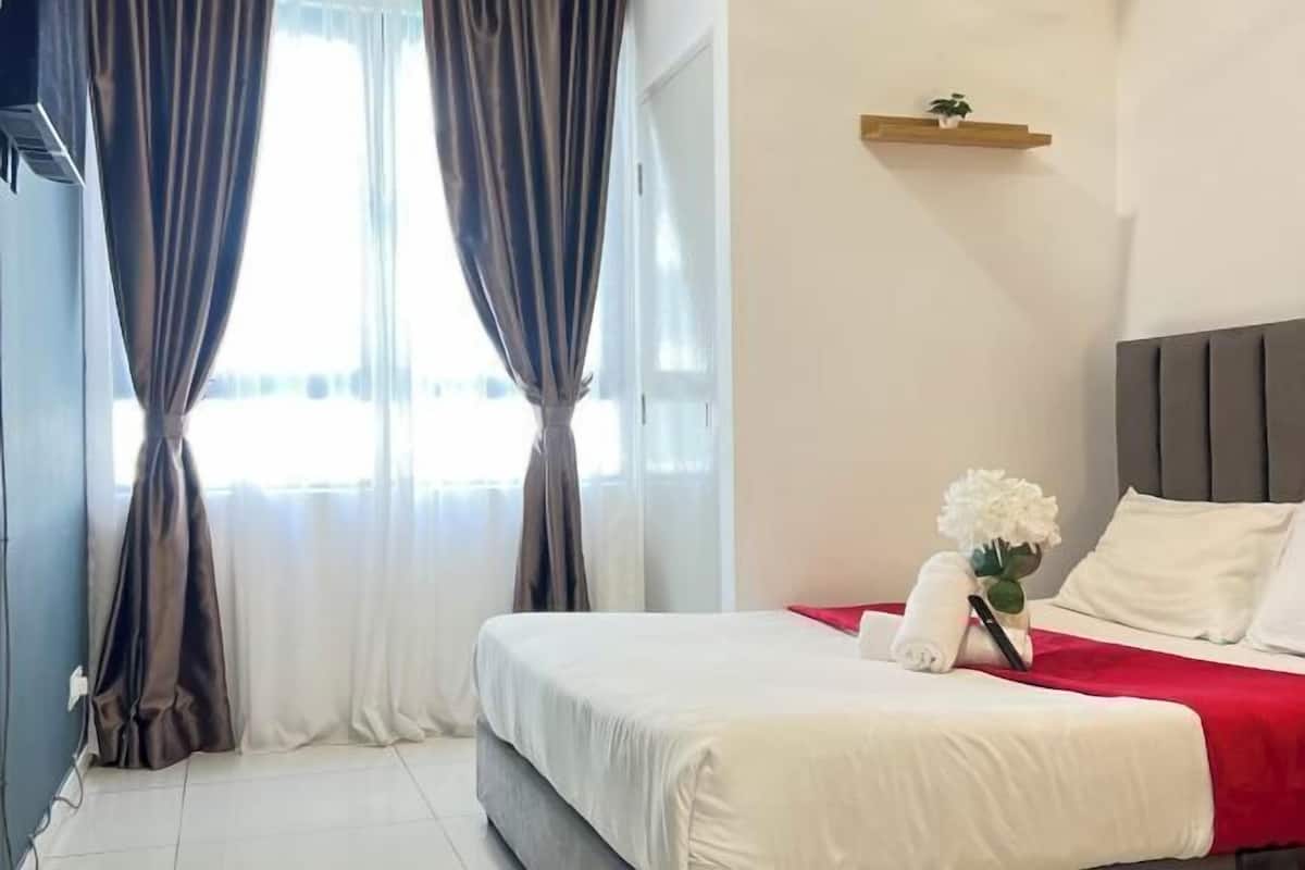 A hotel room with a bed, curtains, a bedside table with a vase of flowers, and a shelf with a plant.