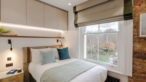 A bedroom with a bed, bedside table, lamp, window, and view of the outside. - Kensington Cosy Studio (London)