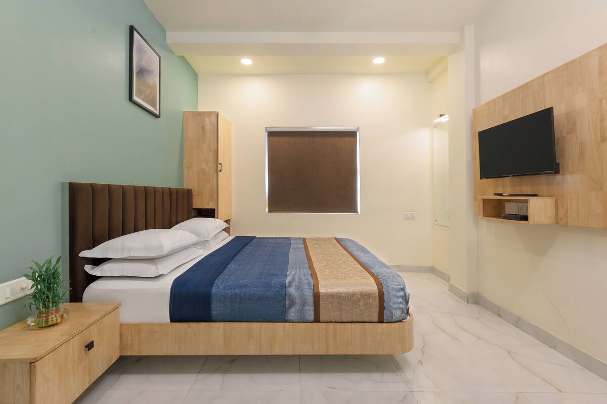 A modern bedroom with a wooden bed, a TV mounted on the wall, and a window with blinds.
